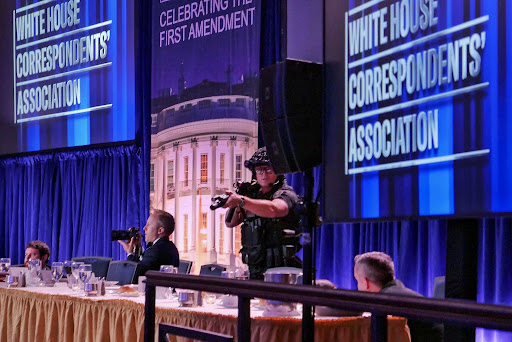 This photo shows a long table with yellow crimping on the side at the front of the image on the left side, while on the right, a metal bar blocks the way. There are many glasses on the table, with two people crouched at each end, while a cameraman is in the middle and a standing officer with a weapon in the center. In the top right and left of the image, two screens read” White House Correspondents’ Association’. 